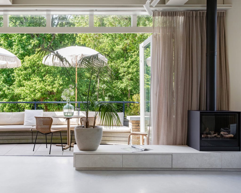 A bright living room with large glass sliding doors overlooking a green environment and a balcony with a sofa, table, and plants.