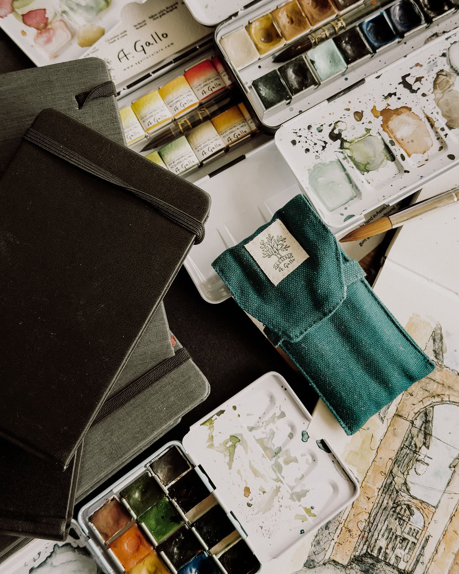 Boxes of watercolor paint, sketchbooks in various sizes, and a paintbrush.