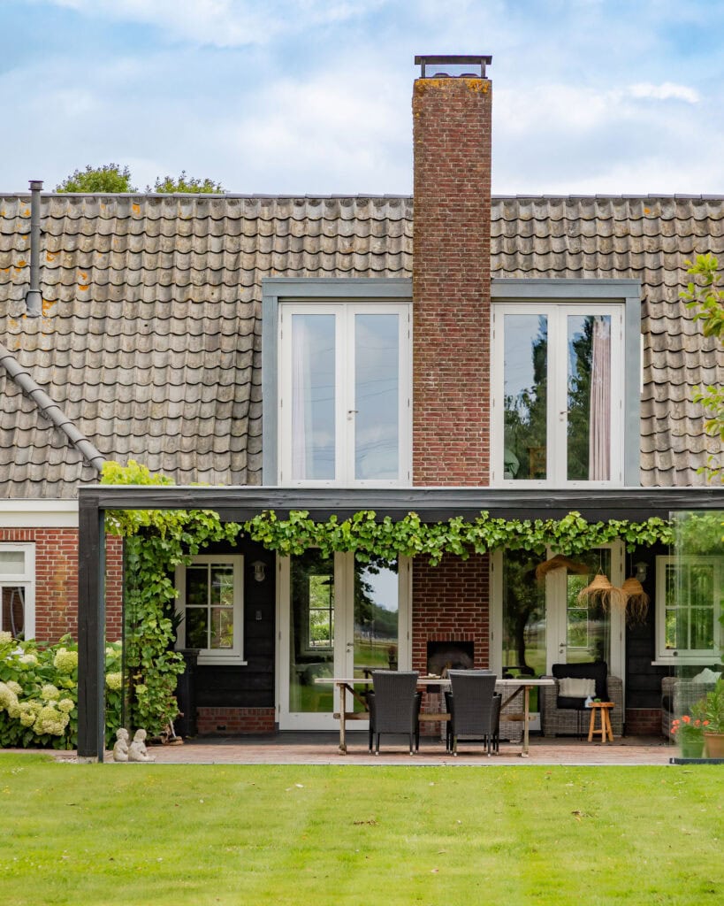 A house with large windows and a terrace in the garden, surrounded by green plants and a well-maintained lawn.