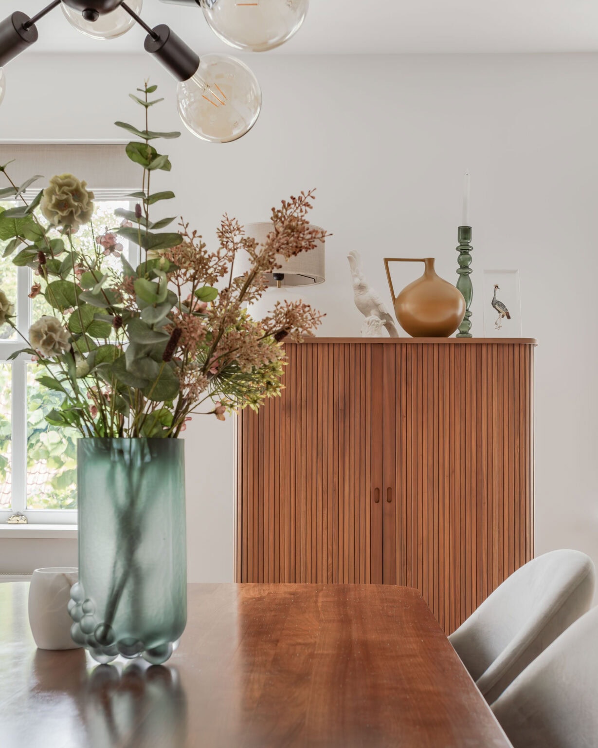 Een stijlvolle interieurfoto met een grote vaas met bloemen op een houten tafel, een houten kast en decoratieve objecten in een lichte woonkamer.