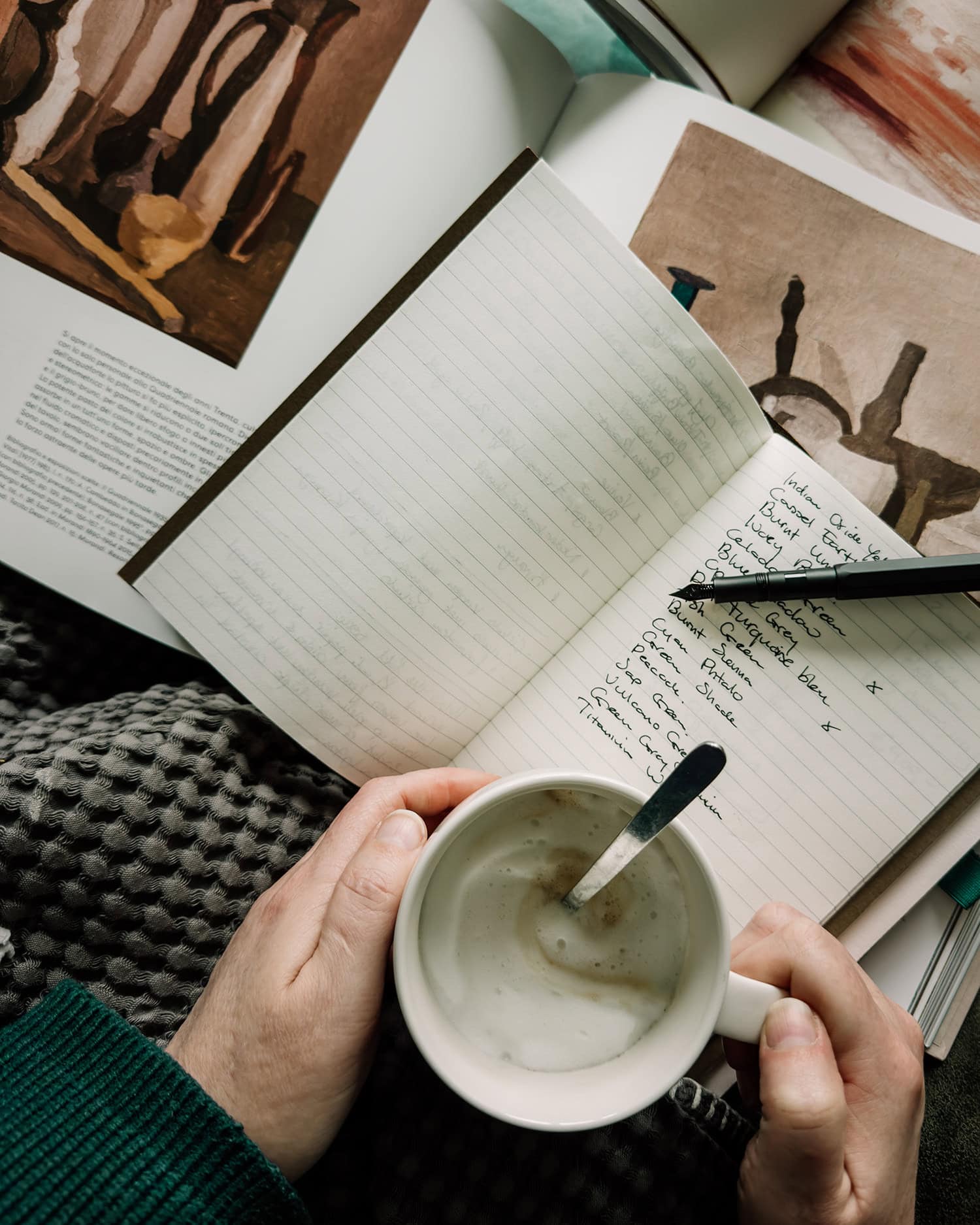 Hands holding a cup of cappuccino, a notebook with a fountain pen showing written paint colors, and art books in the background.