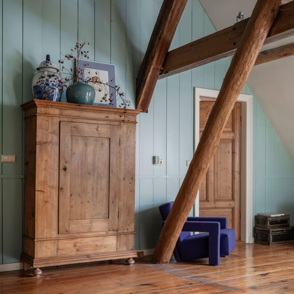 Wooden cabinet in a stolp farmhouse with a blue armchair. The wall is light blue and the square support posts are visible.