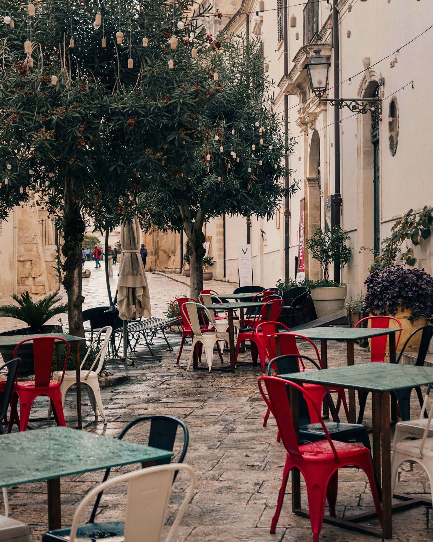 A terrace with red and white chairs and small green tables under the trees in Scicli, Sicily.