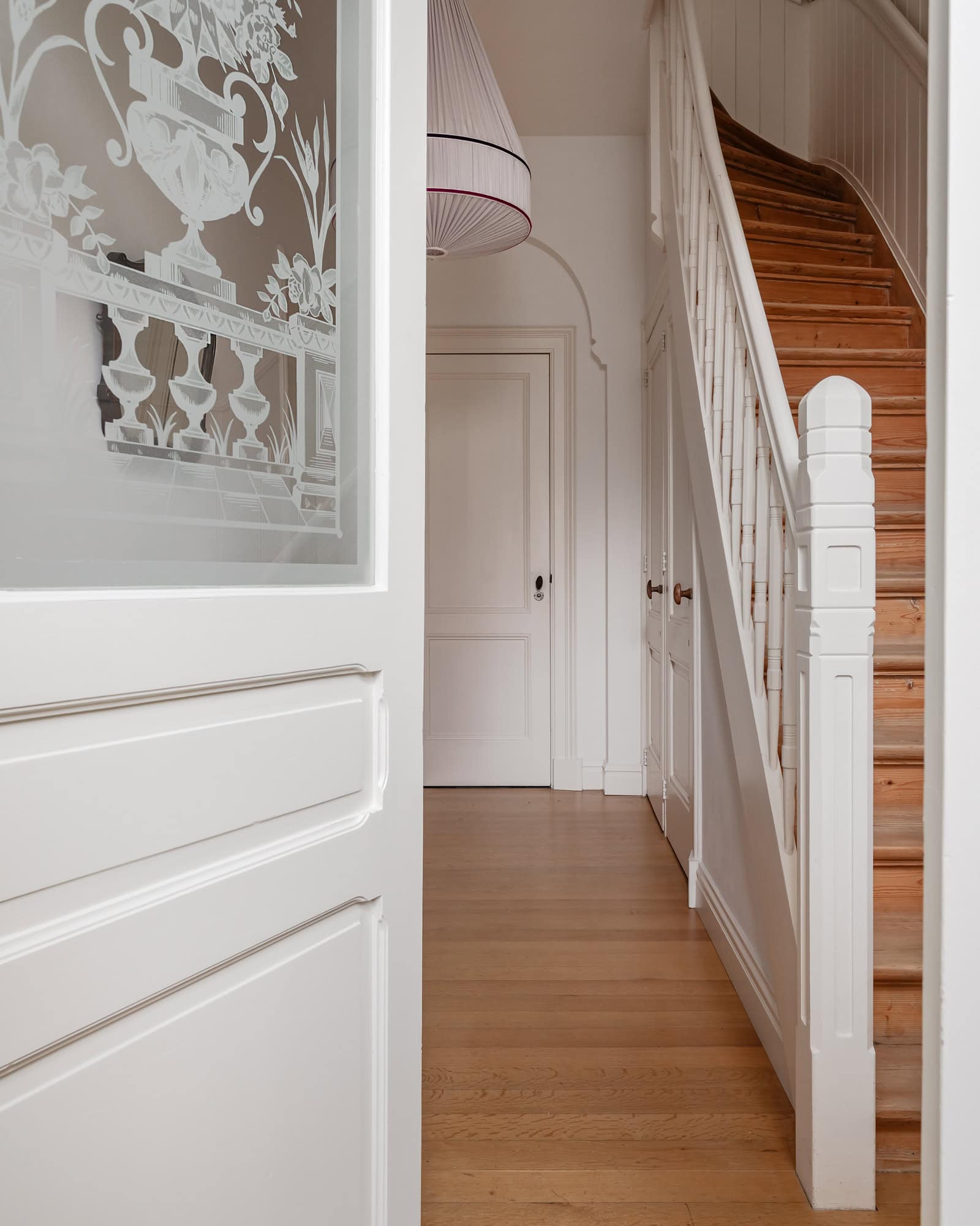 A view through an old door with beveled glass, looking toward a hallway with a wooden staircase.