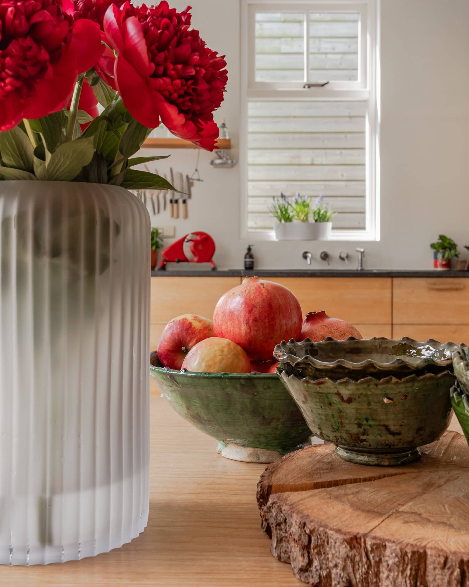 An oak kitchen with a soapstone countertop and red accessories. In the foreground, a vase with red peonies and green bowls.