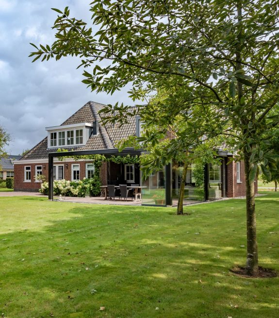 Een modern huis met grote ramen en een terras, omgeven door een groene tuin en bomen.