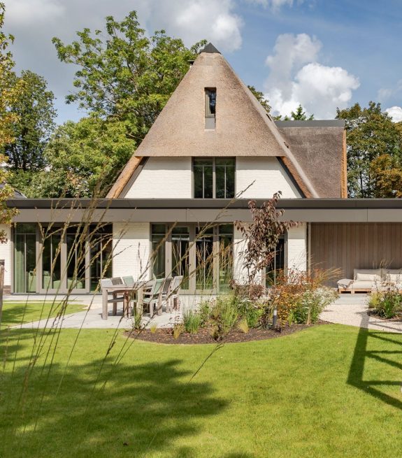 Rietgedekte villa met grasveld op de voorgrond en bomen op de achtergrond.